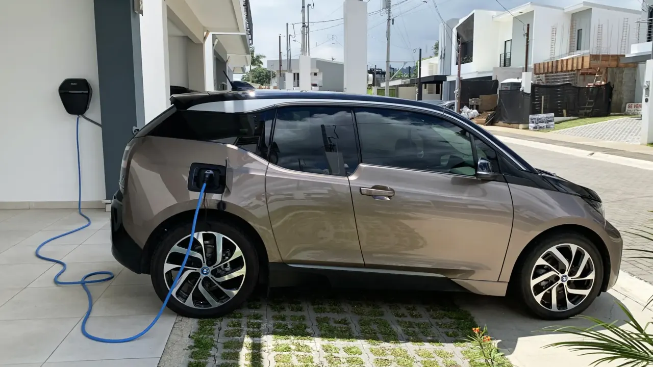 Carregar carro elétrico em casa: BMW i3 marrom carregando na garagem de casa com cabo azul ligado a um wallbox, ilustrando…