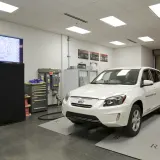 Carro elétrico branco sobre placas de recarga sem fio em movimento durante demonstração em laboratório, com tela de monitor…