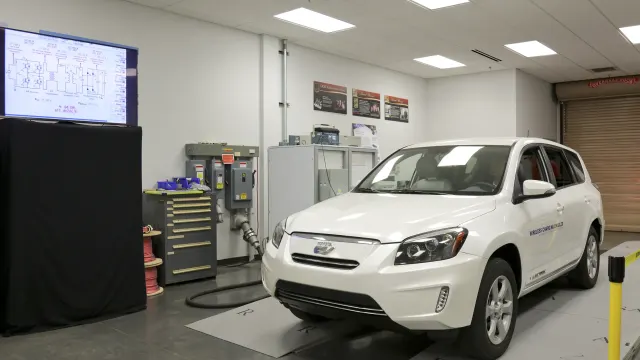 Carro elétrico branco sobre placas de recarga sem fio em movimento durante demonstração em laboratório, com tela de monitor…